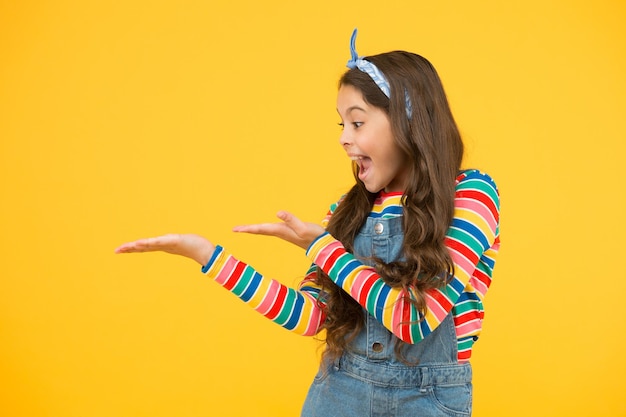 Girl demonstrate product. Promoting product for kids. Launching product. Advertisement concept. Product presentation. Kid happy smiling face show something on open palm copy space yellow background.