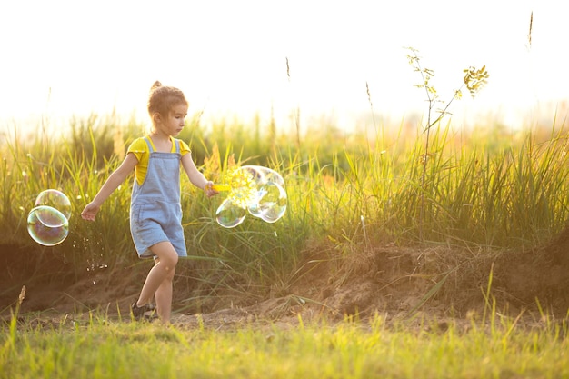 A girl in a denim jumpsuit blows soap bubbles in the summer in a field at sunset. International Children's Day, happy child, outdoor activities. Summer background. Healthy and eco-friendly lifestyle