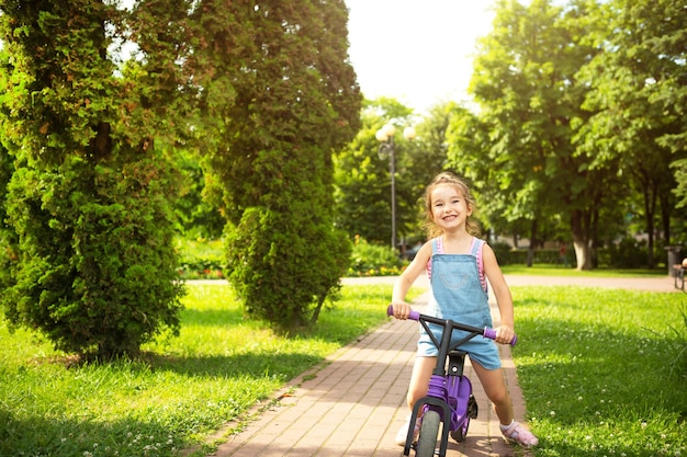 A girl in a denim jumpsuit rides a purple running bike in the summer