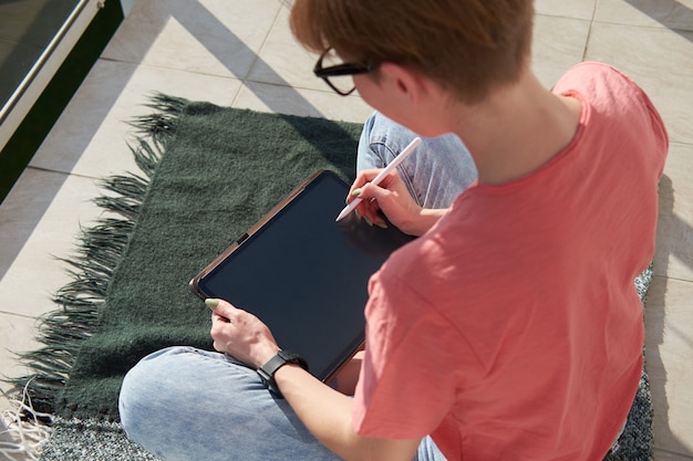 Girl digital artist drawing on tablet while sitting on sunny balcony. Freelance work