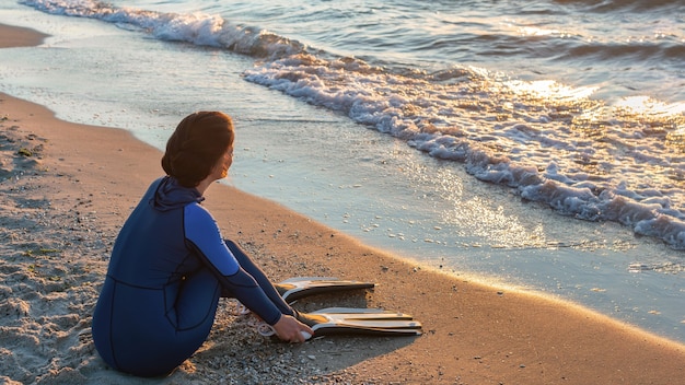 A girl diver sits on the sea shore and puts on her fins