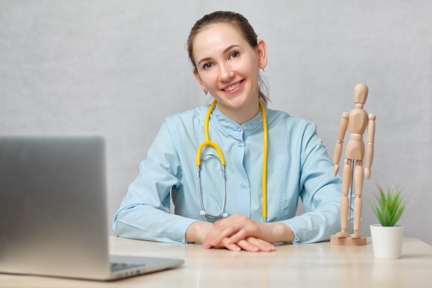 Terapista medico ragazza si siede a un tavolo con un computer portatile su uno sfondo bianco e guarda la telecamera.