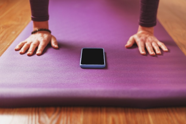 A girl does yoga with a smartphone online via the Internet on a lilac rug in the living room.