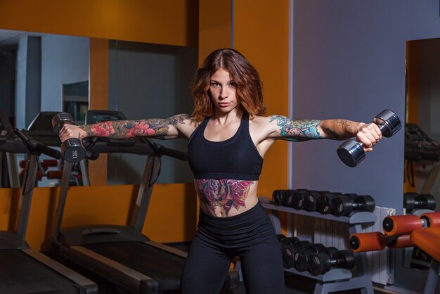 Ragazza che fa esercizi con manubri. la ragazza con i tatuaggi sul fitness dimostra un bel corpo con i muscoli. varie attrezzature sportive e attrezzature per il fitness per lo sport e il fitness