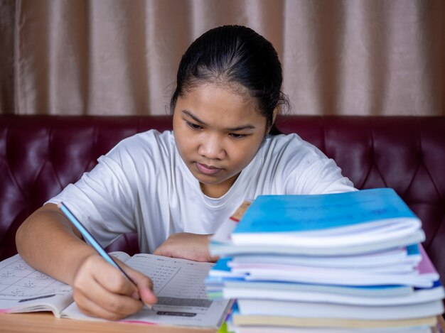 Girl doing homework on a wooden table and there was a pile of books next to it The background is a red sofa and cream curtains.