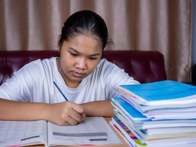 Girl doing homework on a wooden table and there was a pile of books next to it The background is a red sofa and cream curtains.