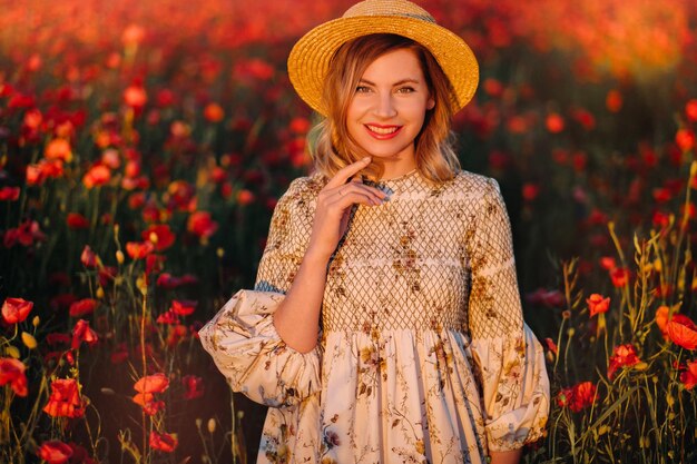 A girl in a dress in a hat walks in a field with poppies at sunset