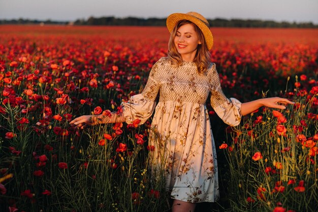 A girl in a dress in a hat walks in a field with poppies at sunset