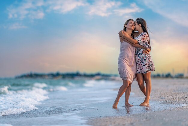 A girl in a dress hugs her mom in a dress while walking along the beach near the sea with waves