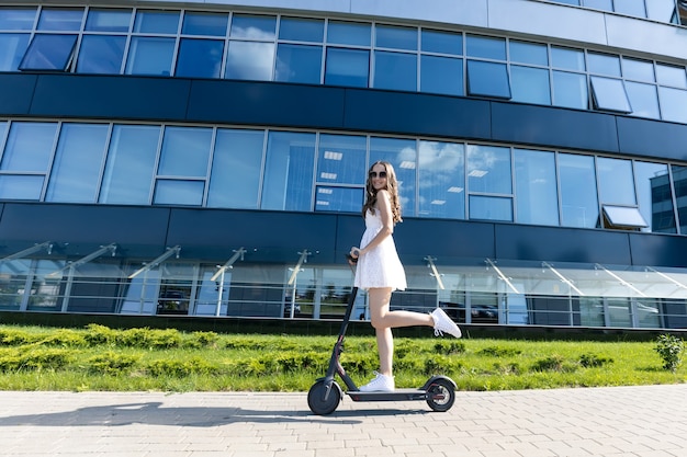 A girl in a dress rides an electric scooter near modern buildings