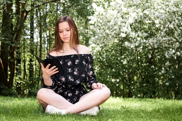 Girl in a dress sits on the grass in the park and reads an e-book