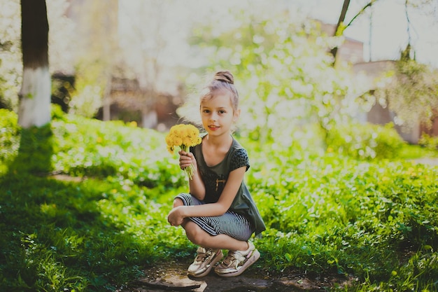 ドレスを着た女の子が春の桜の庭で黄色いタンポポの花束を嗅ぎます