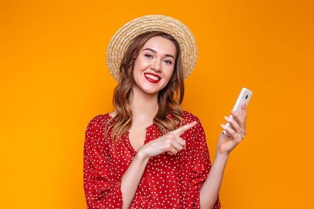 Girl in a dress, a straw hat holds a mobile phone and points a finger at him isolated over orange background