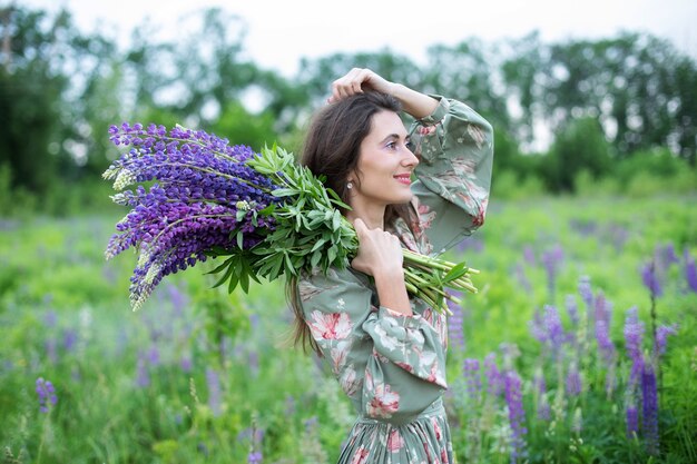 A girl in dress with lupine flowers stands in field girl with bouquet of wild flowers