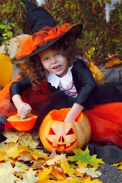 주황색 치마에 작은 마녀로 옷을 입고 뾰족한 검은 모자를 입은 소녀가 가을 공원에서 할로윈 호박 옆에 앉아 있습니다.