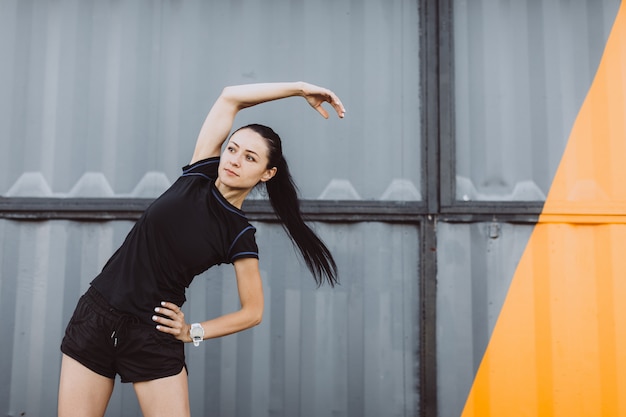 黒のTシャツとショートパンツを着て、黄色の灰色の壁で実行する前にウォーミングアップする女の子