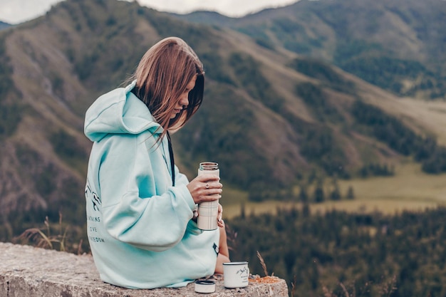 A girl dressed in a blue hoodie with a print is sitting on a
stone and is about to pour a hot drink from theromos for traveling
into a mug. background of rocky hills.