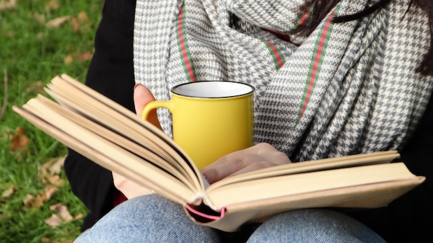 A girl dressed in a coat and a scarf in the autumn forest holds a book and a cup with a hot drink in her hands close-up in a city park on a warm day. The concept of reading, relaxation and comfort.