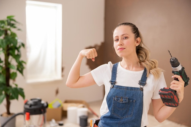 A girl dressed in denim style holds a drill in her left hand and shows her biceps with right hand