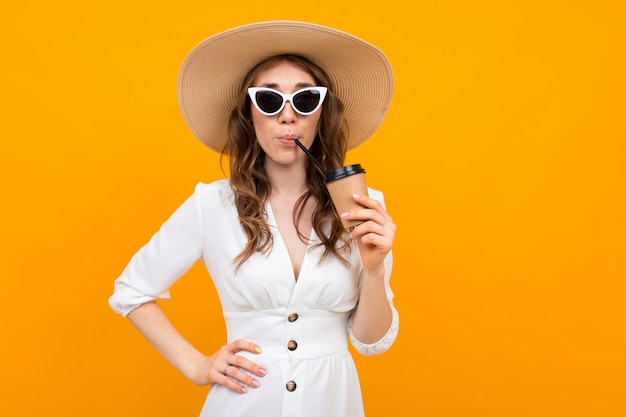 Girl dressed in a white dress with a hat i in glasses drinks a drink on a yellow background
