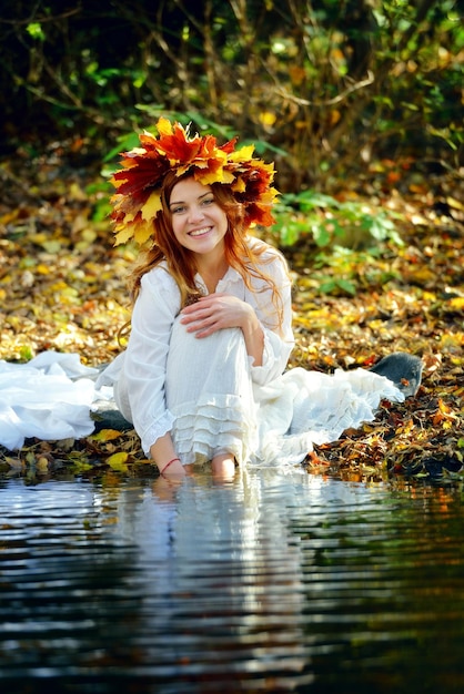 A girl dressed in white wearing a wreath of autumn leaves sits by a stream on a sunny day