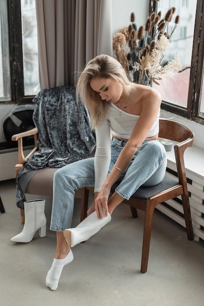 Девушка одевает белые кожаные туфли Красивая молодая блондинка в модной повседневной одежде сидит на стуле и надевает обувь в комнате