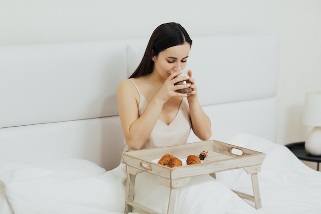 Ragazza che beve cappuccino e mangiare la colazione sul letto al mattino