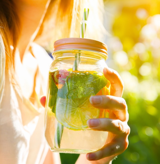 ストローで瓶に新鮮なレモネードを飲む女の子。流行に敏感な夏の飲み物。自然の中で環境にやさしい。ガラスにミントが入ったレモン、オレンジ、ベリー。