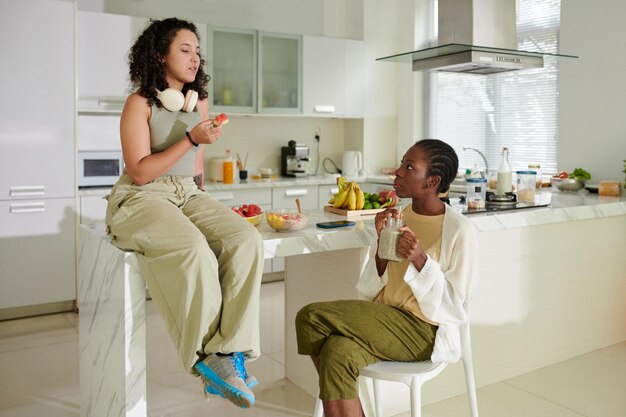 Girl drinking smoothie and looking at friend eating watermelon and telling news