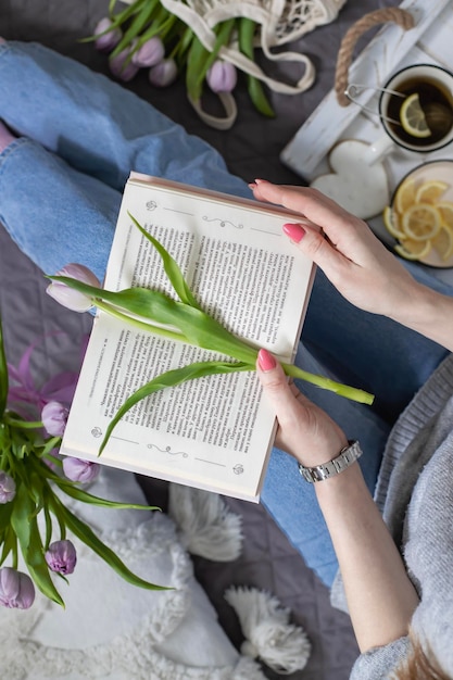 The girl drinks tea and reads a book while sitting on the bed