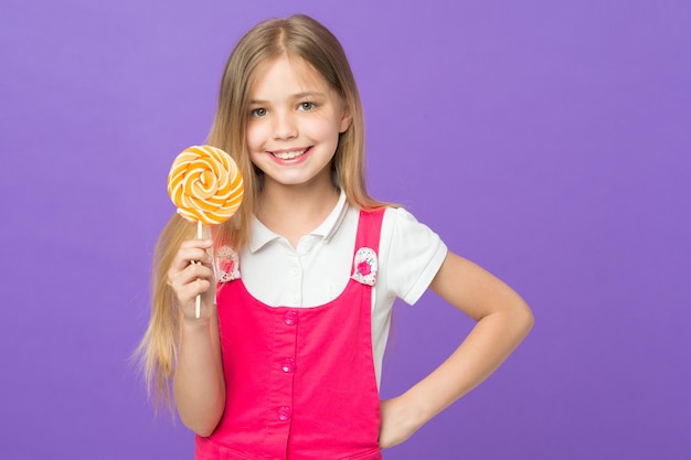 Girl eating big candy on stick or lollipop sweet childhood concept kid with long hair likes sweets and treats girl on smiling face holds giant colorful lollipop in hand violet background