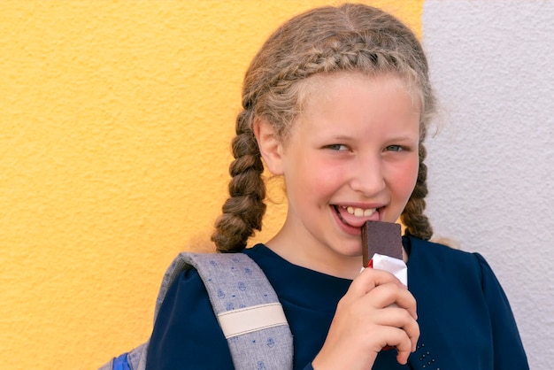 チョコレートバーのクローズアップを食べる女の子。背景の女子高生の肖像画。コンセプトの学校給食。チョコレートバーを持つ女の子。菓子の棒を持つ生徒。コピースペース。コピースペース