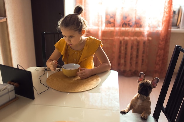 Girl eating at the kitchen and watching video on tablet home lifestyle portrait dog on floor