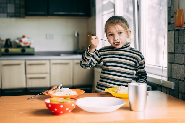 縞模様のジャケットのキッチンでソーセージとパスタを食べる女の子