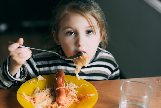 Photo girl eating pasta with sausage in the kitchen in a striped jacket