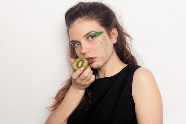 Photo the girl eats a half kiwi. happy young beautiful ecstatic beautiful young woman holding two cut kiwis on her eyes for fun joke or dynamic vitamin c, indoors