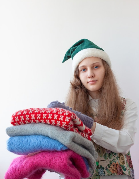 A girl in an elf hat holds several knitted christmas sweaters in her hands.