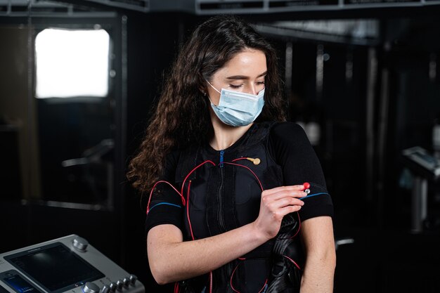 Girl in EMS suit and medical mask in gym. Protection from coronavirus covid-19. Sport training in electrical muscle stimulation suit.