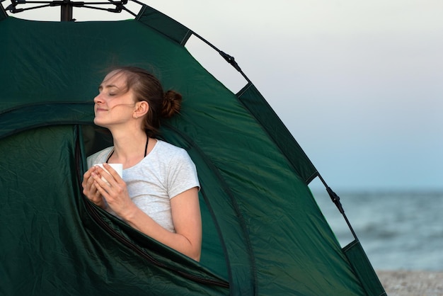 Girl enjoys the morning in a tent by the sea Mug of tea in hands Travel hiking camping