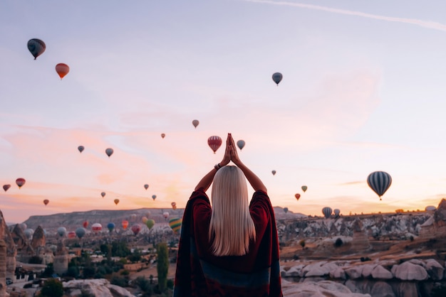 Girl in ethnic clothes at dawn watching the flight a lot of balloons fly over the valley of love