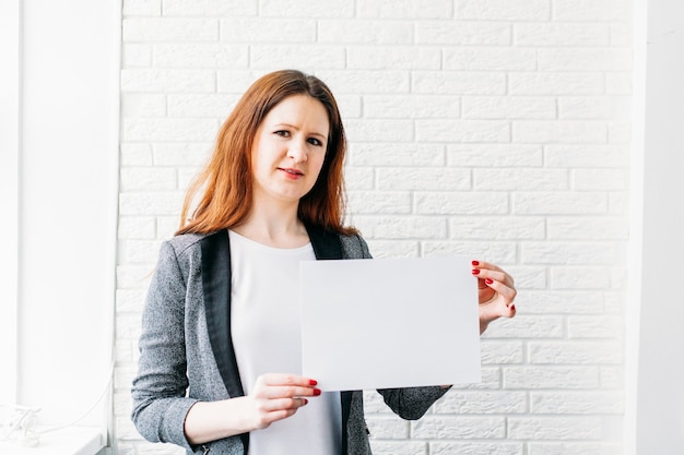 A girl of European appearance holds a white sheet of paper for the placement of an inscription, advertisement, slogan, trademark