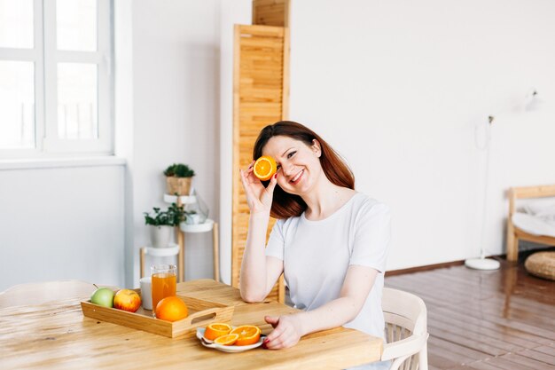 Photo a girl of european appearance sits at a table in a white t-shirt, oranges for making smoothies and juice, the benefits of vitamins, morning