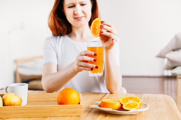 A girl of European appearance sits at a table in a white T-shirt, oranges for making smoothies and juice, the benefits of vitamins, morning