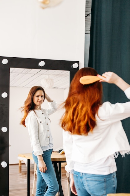 Photo a girl of european appearance stands in front of a mirror, combs her hair with a comb, hair beauty, care,