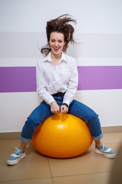 A girl of European appearance with different emotions on her face sits on a ball for a gymnast. Attractive young brunette woman smiling and fooling around