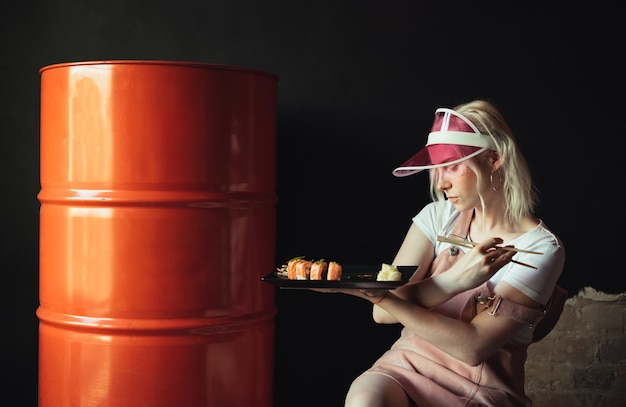 girl in fancy pink clothes posing on a barrel indoors with a plate of sushi rolls in her hands