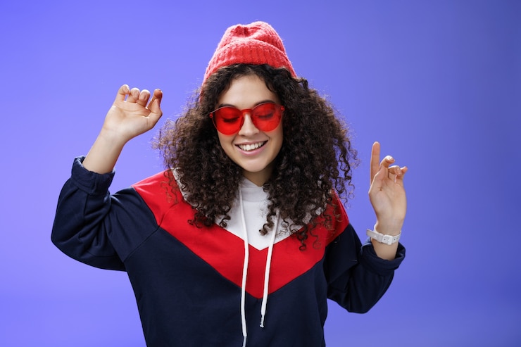 Photo girl feeling awesome having fun and enoying cool party, dancing with raised hands looking down smiling broadly wearing sunglasses and stylish beanie posing against blue background.