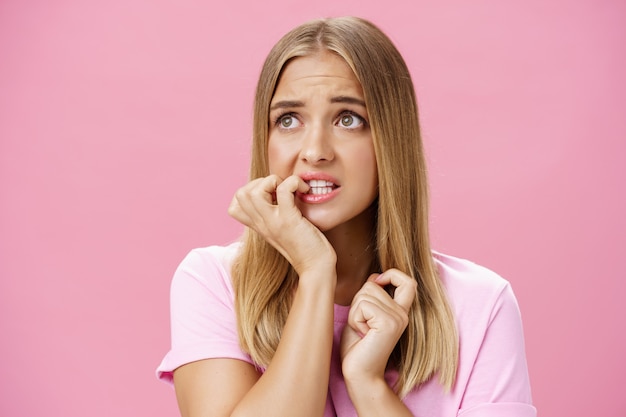 Girl feeling concerned and scared looking aside with anxiety and panic biting fingernails from stress and troublesome situation posing over pink wall