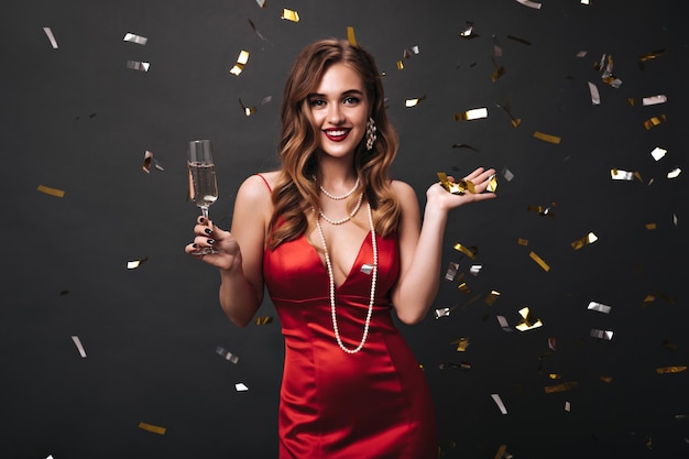 Girl in festive outfit and jewelry holding champagne glass Pretty happy woman in red silk dress drinking white wine on black background with golden confetti