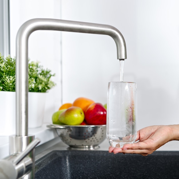 The girl fills a glass with water from a water tap in the kitchen.